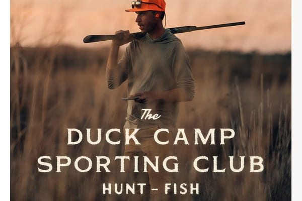 A hunter in Duck Camp gear standing in a marsh at sunrise, embodying the wingshooting lifestyle.